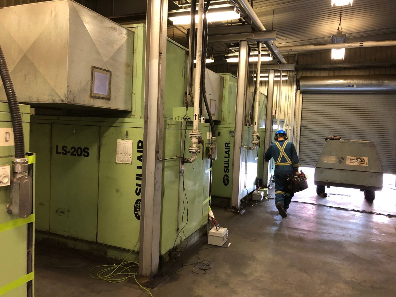 Worker in a safety harness and helmet walking through an industrial facility next to large green Sullair air compressors.