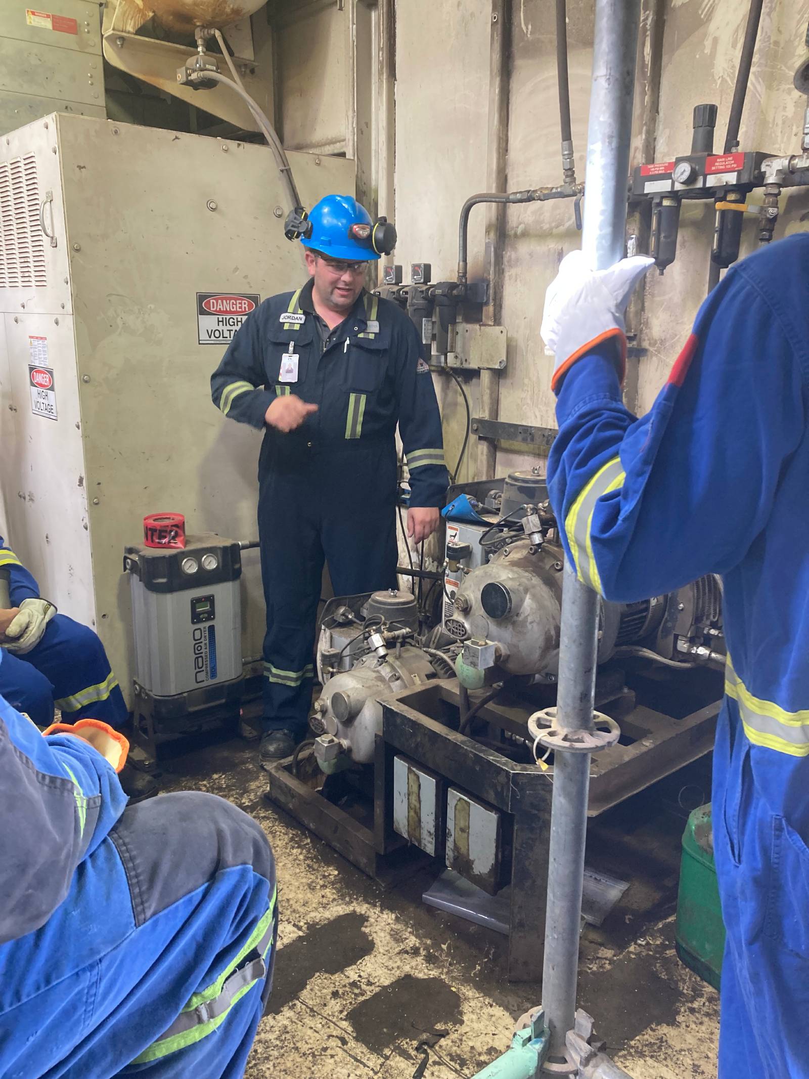 A technician in a blue hard hat and safety gear standing with other workers surrounding shovel compressors in an industrial setting.