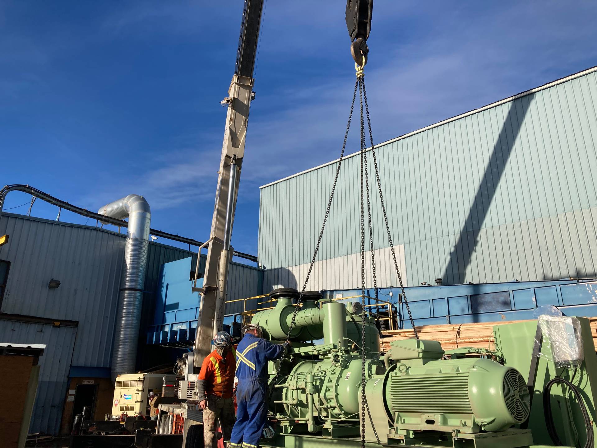 Two workers guide the installation of a large Sullair oil-flooded air compressor using a crane outside an industrial facility.