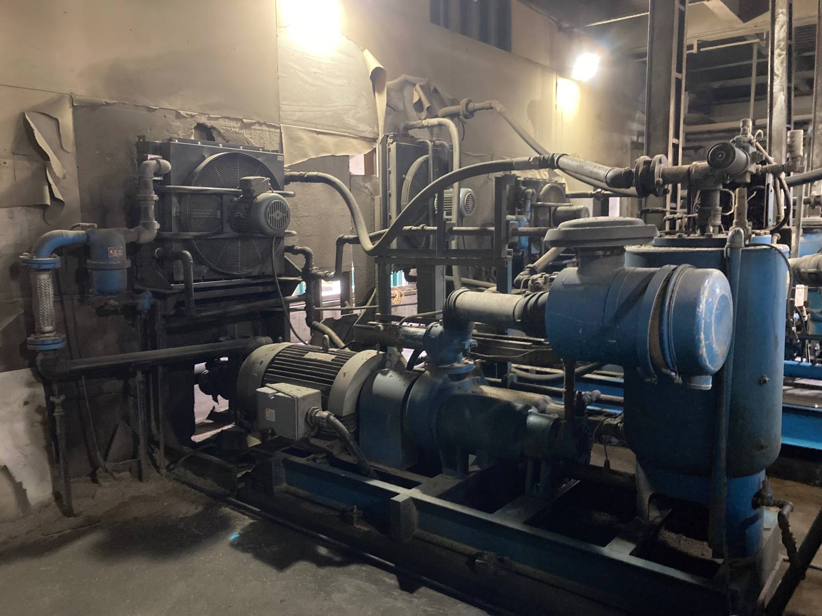 Original-Sawmill-Equipment Old industrial compressor system with worn blue components and visible signs of wear in a dimly lit mechanical room.
