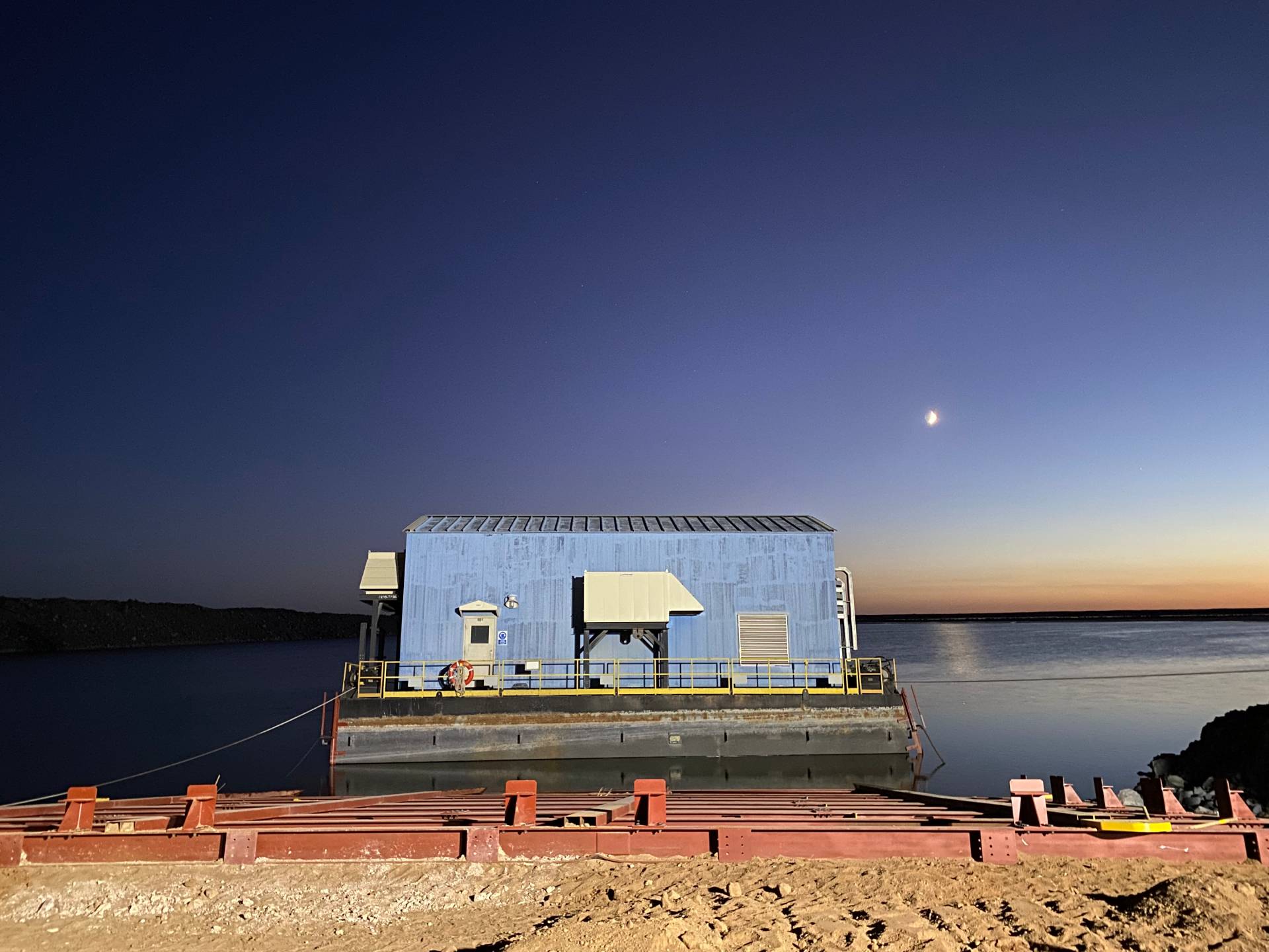 A barge on calm water in a remote Mongolian mine, with a crescent moon visible in the sky during the relocation project.