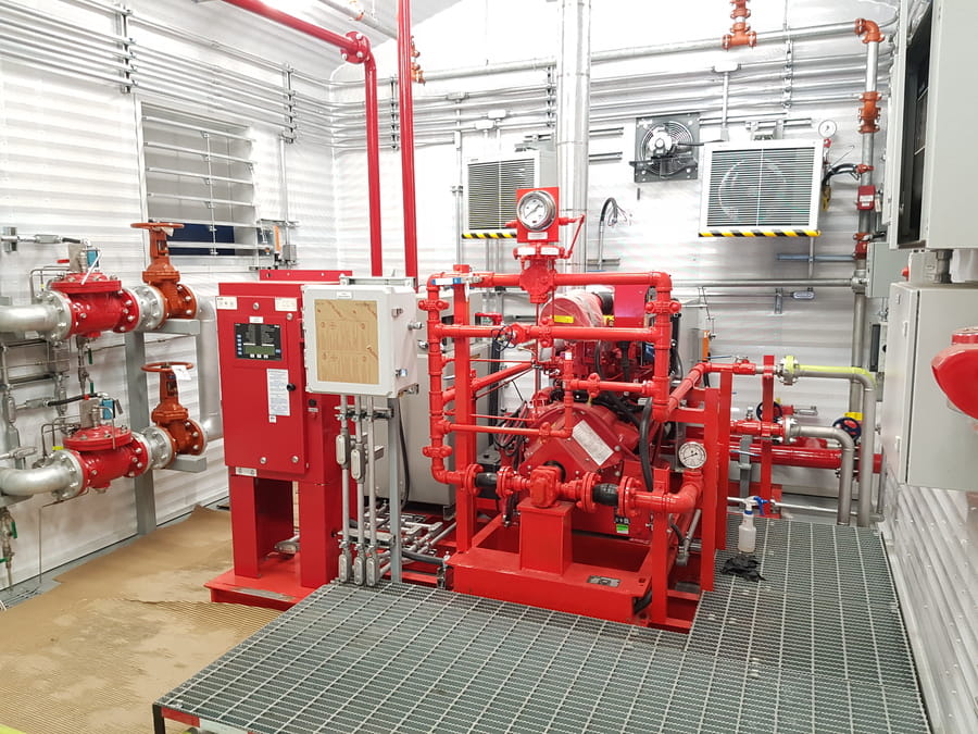 A brightly lit mechanical room features red industrial equipment, including gauges, valves, and piping. Metallic walls and fans add a technical, sterile vibe.
