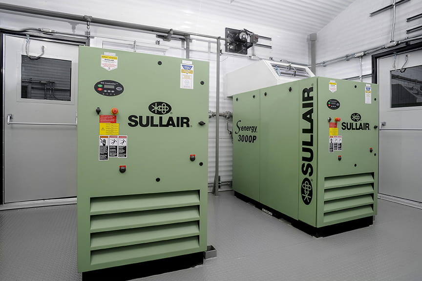 Green Sullair air compressors in a clean, industrial room with metal walls and doors. The setting is orderly, showcasing modern equipment.