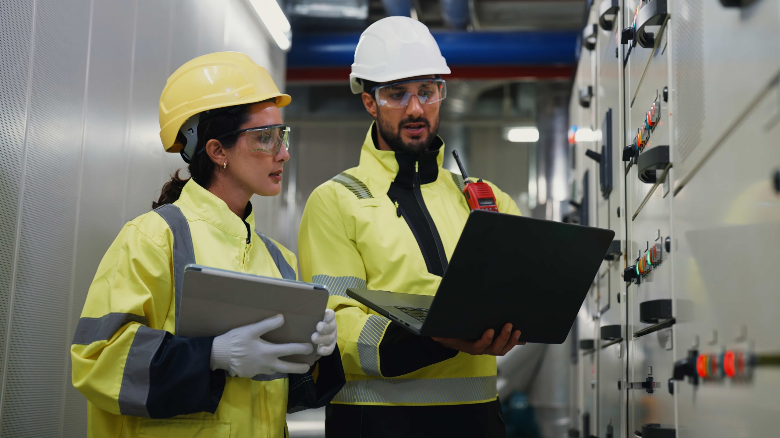Two engineers in hi-vis safety jackets and hard hats reviewing equipment data on a laptop and tablet in front of an industrial electrical control panel